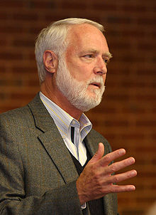 A white-haired and white-bearded man gesturing with his right hand as he speaks