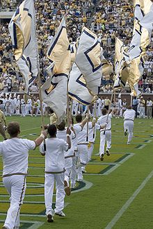 Eight men dressed in white uniforms run in a line across the stadium endzone carrying large white and gold flags.