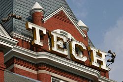 Large, white, capital letters spelling "TECH" situated just below the pointed roof of the square, red brick tower of the administration building
