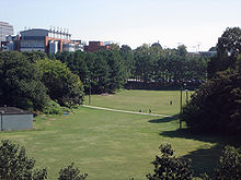 A grassy field surrounded by large, green-leafed trees and bushes. A sidewalk cuts through the field horizontally. A large red brick and white concrete building with idealized smoke stacks is in the background to the left, as are a few other less visible buildings.